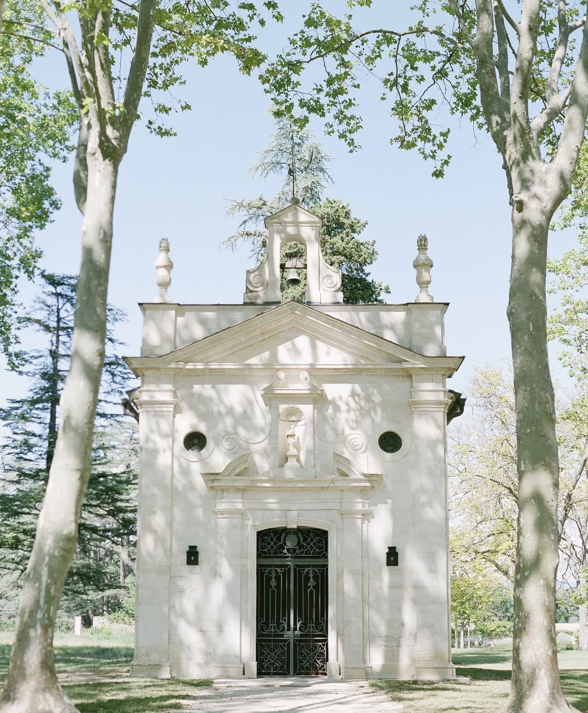 A small white neoclassical Catholic chapel with a pedimented stone façade, bell tower, and ornate wrought-iron doors, framed by tall plane trees in dappled sunlight.